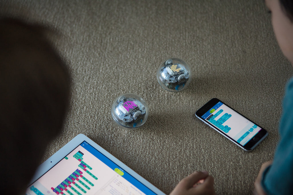 Two kids use block coding on a tablet and smartphone to code their Sphero BOLT programmable robots.