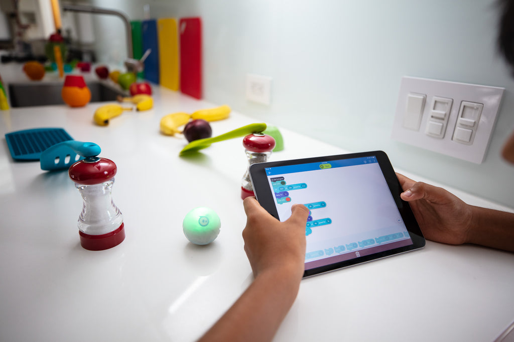 A Sphero Mini is coded through a maze of kitchen supplies by an iPad with block code.