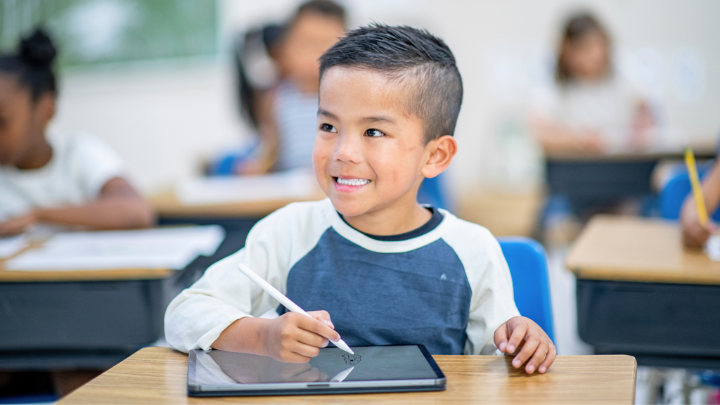 A young boy works on a tablet at a table in his classroom.