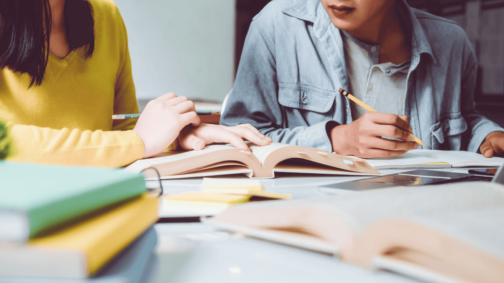 Two female students read from a textbook and take notes with a pencil and paper.