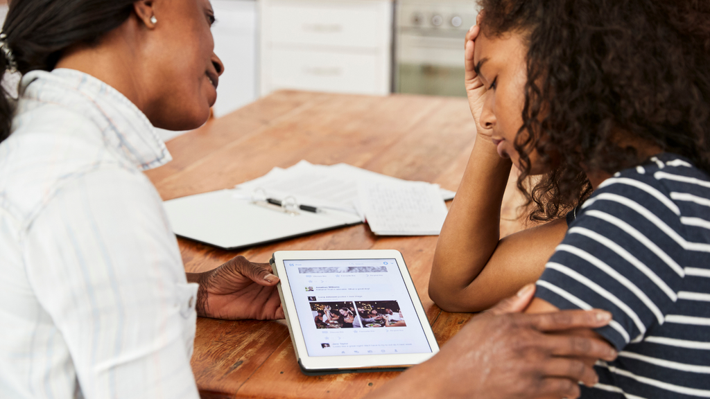 A frustrated girl and her mother sit at the table looking at a lesson on a tablet.