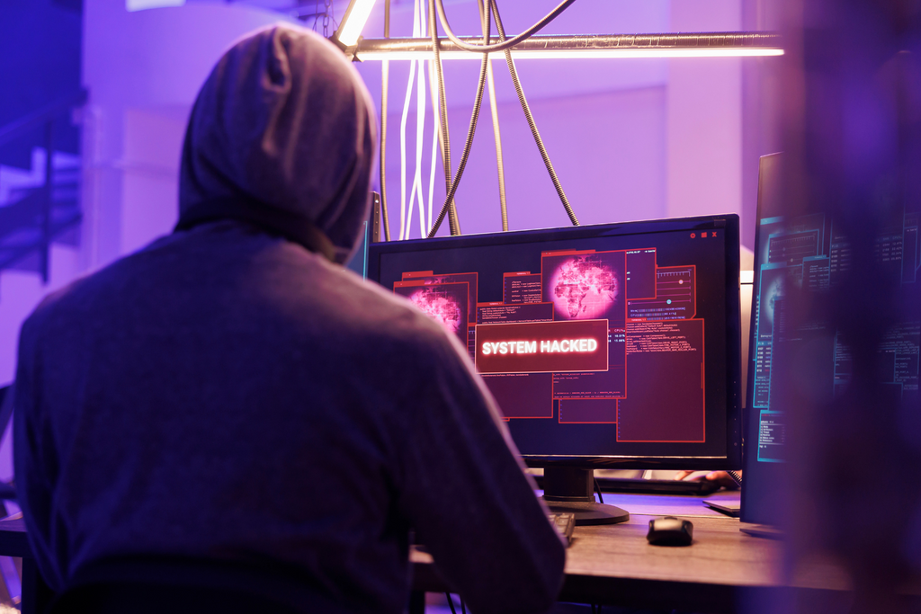 A hooded person working on computer screen reading "system hacked".