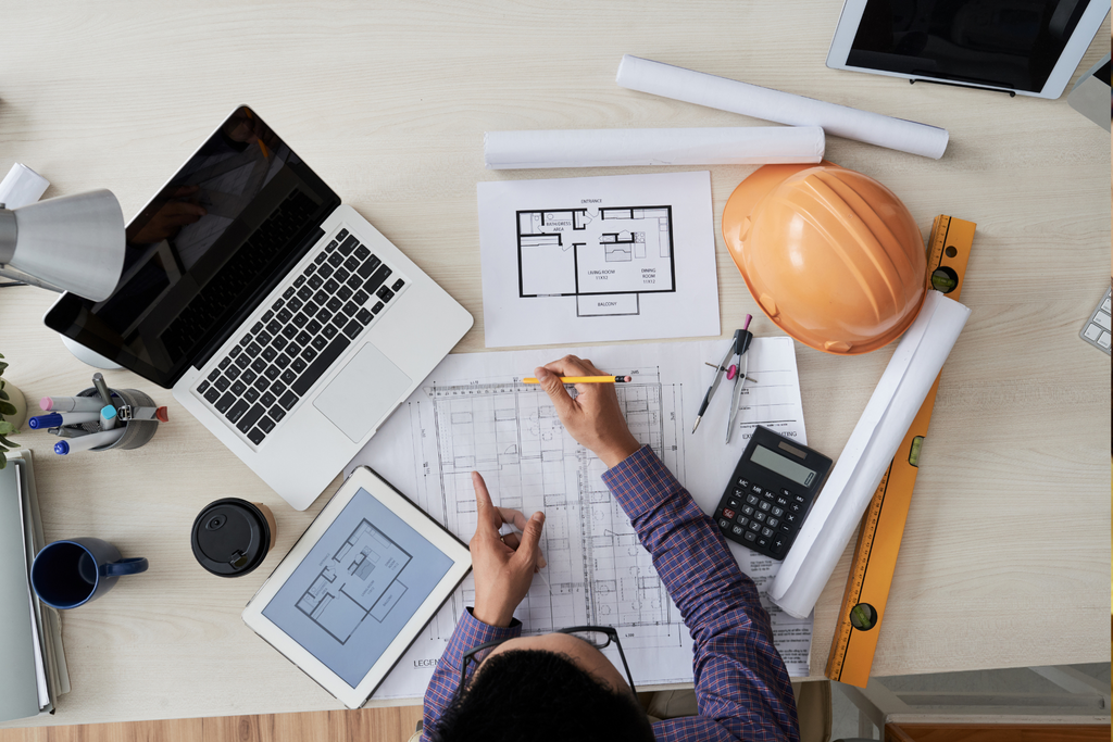 A birds-eye view of a civil engineer working at a desk containing blueprints and other construction tools..