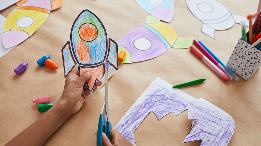 A kids' hands using scissors to cut out a drawing of a rocket ship.